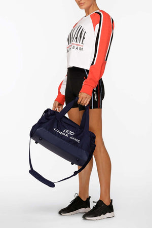 A woman in sporty attire holds the Lorna Jane Essential Gym Bag with an adjustable strap. She wears a long-sleeve cropped top featuring "Lorna Jane," black shorts, and black sneakers, standing against a plain white background.