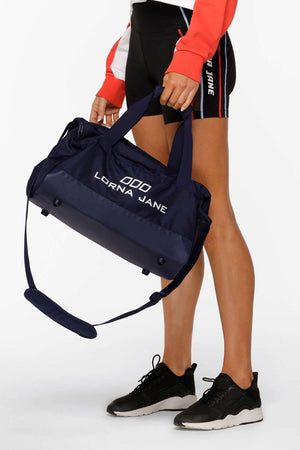 A woman in athletic shorts and sneakers holds the Lorna Jane Essential Gym Bag by its handles, with the adjustable shoulder strap hanging down.