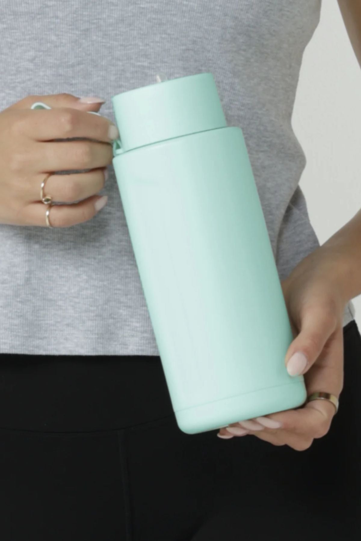 A person in a gray top and black pants holds the Lorna Jane Essential Insulated Water Bottle (1L, light blue) by its handle, gripping it securely with ring-adorned fingers.