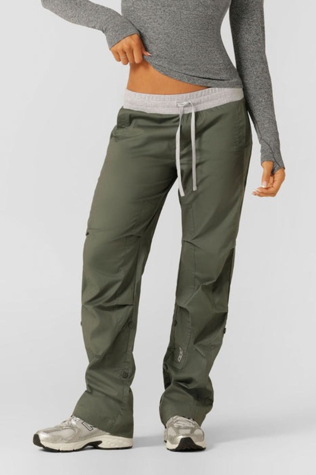 A person wears olive green Lorna Jane Flashdance Pants with an adjustable waist and light grey waistband, paired with a fitted long-sleeve top and white sneakers. The background is plain, and the person's face is not shown.