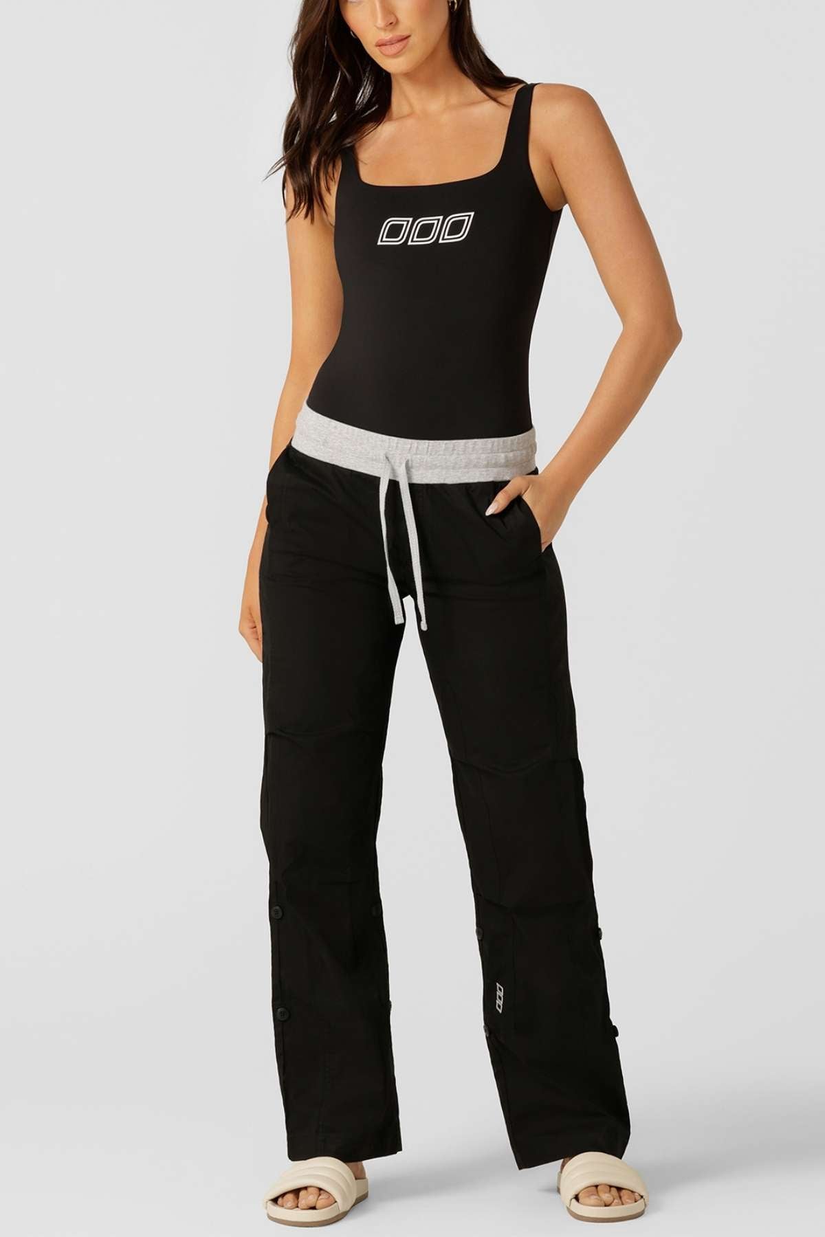 A woman stands with one hand in her pocket, wearing a black tank top with a white graphic, beige slide sandals, and Lorna Jane Flashdance Pant—black cargo pants with an adjustable elastic waist and drawstring.