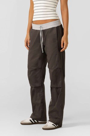 A person wearing a white, sleeveless, striped top and Lorna Jane Flashdance Pant in dark gray with reinforced seams and white sneakers stands against a plain light gray background.