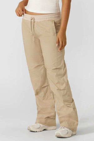 A person models the Lorna Jane Flashdance Pant, featuring a beige multi-pocket cargo style, paired with a white top and white sneakers, standing against a plain light gray background. Only the body from mid-torso to feet is shown.