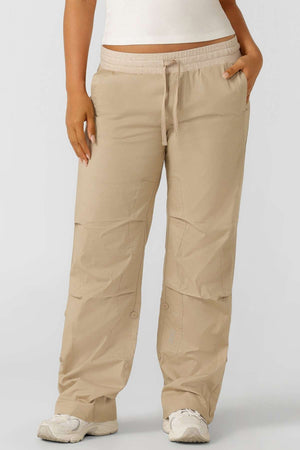 Someone stands against a plain background, wearing a white top, beige winter Lorna Jane Flashdance Pant, and white sneakers, with one hand in their pocket.