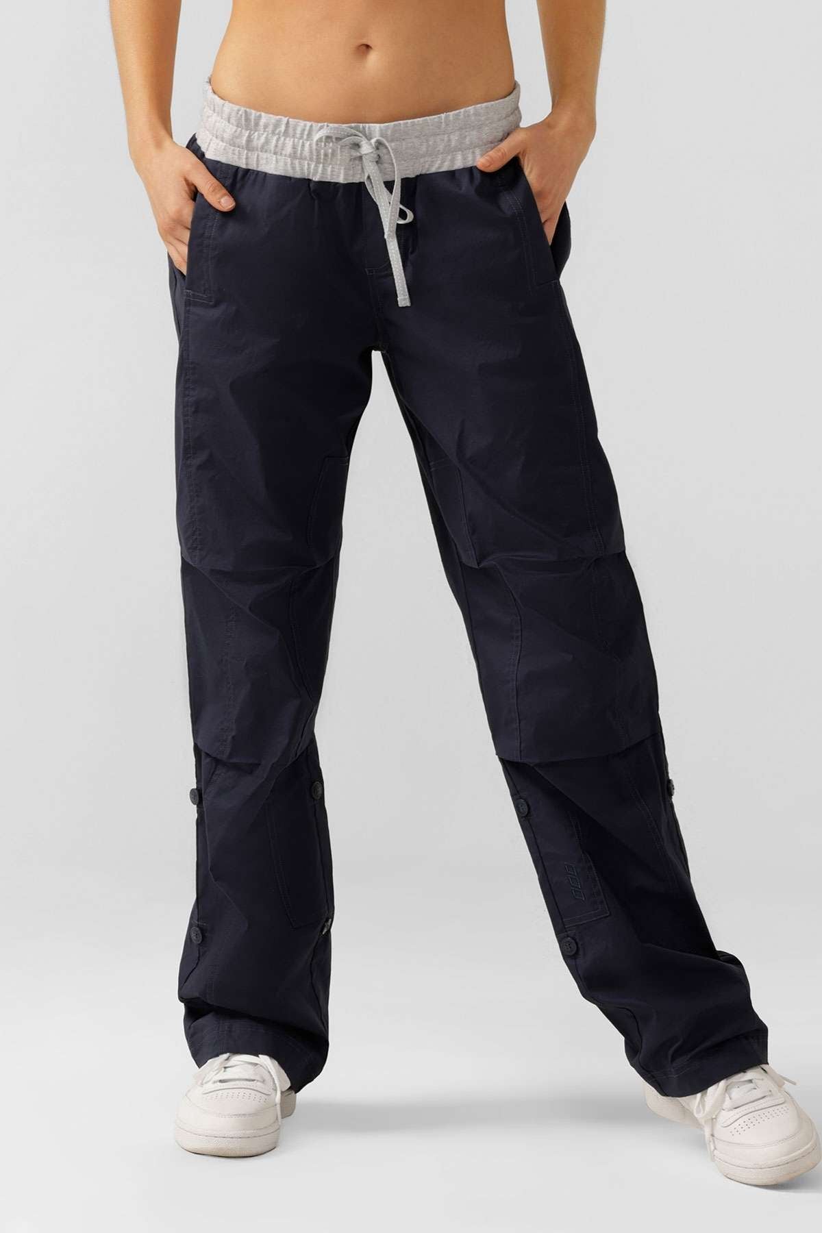 A person wearing loose-fitting black Lorna Jane Flashdance Pants with reinforced seams, a gray drawstring waist, and white sneakers stands with hands in pockets against a plain background.
