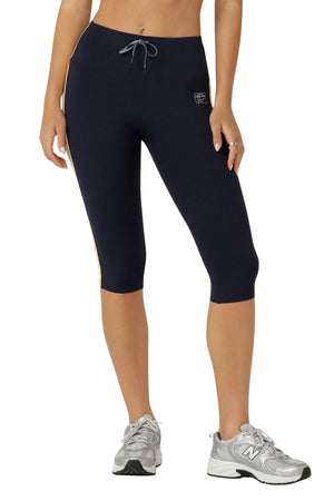 A person wears Lorna Jane Game Day 3/4 Capri Leggings featuring a drawstring waist and small left-hip logo, paired with silver New Balance sneakers, creating a retro sport look against a plain white background.