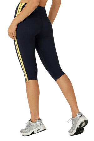A person showcases the Lorna Jane Game Day 3/4 Capri Leggings—navy blue with yellow side stripes in a retro sporty style—paired with gray athletic shoes, standing sideways with one leg bent to reveal the back and side view.