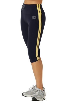 Wearing Lorna Jane Game Day 3/4 Capri Leggings in navy with yellow side stripes and gray athletic sneakers, a person stands on a white background, showcasing a retro sport style.