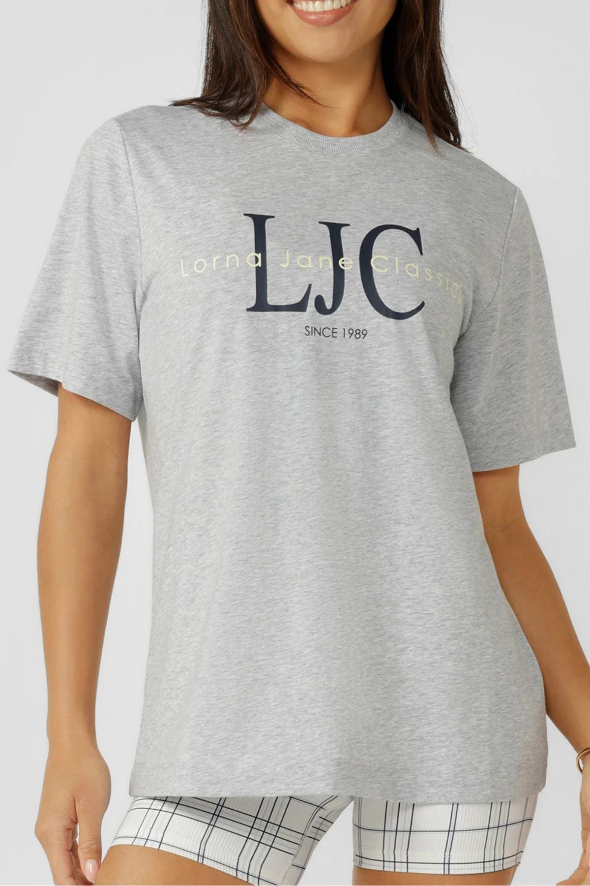 A woman smiles against a neutral background wearing the Lorna Jane Heritage Classics Relaxed T-Shirt in light gray with "LJC Lorna Jane Classics SINCE 1989" on the front, paired with white and black plaid shorts.