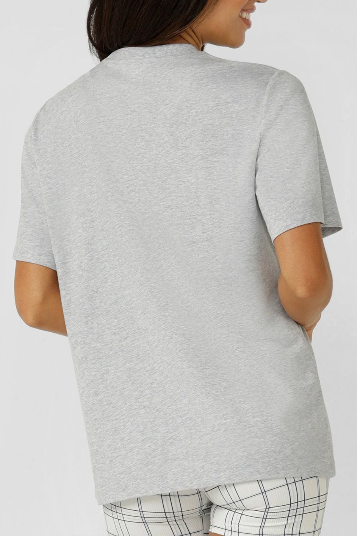 A woman, smiling with one hand in her pocket, wears the Lorna Jane Heritage Classics Relaxed T-Shirt in light gray and plaid shorts. The breathable cotton blend adds comfort, and the plain white background keeps the look fresh.