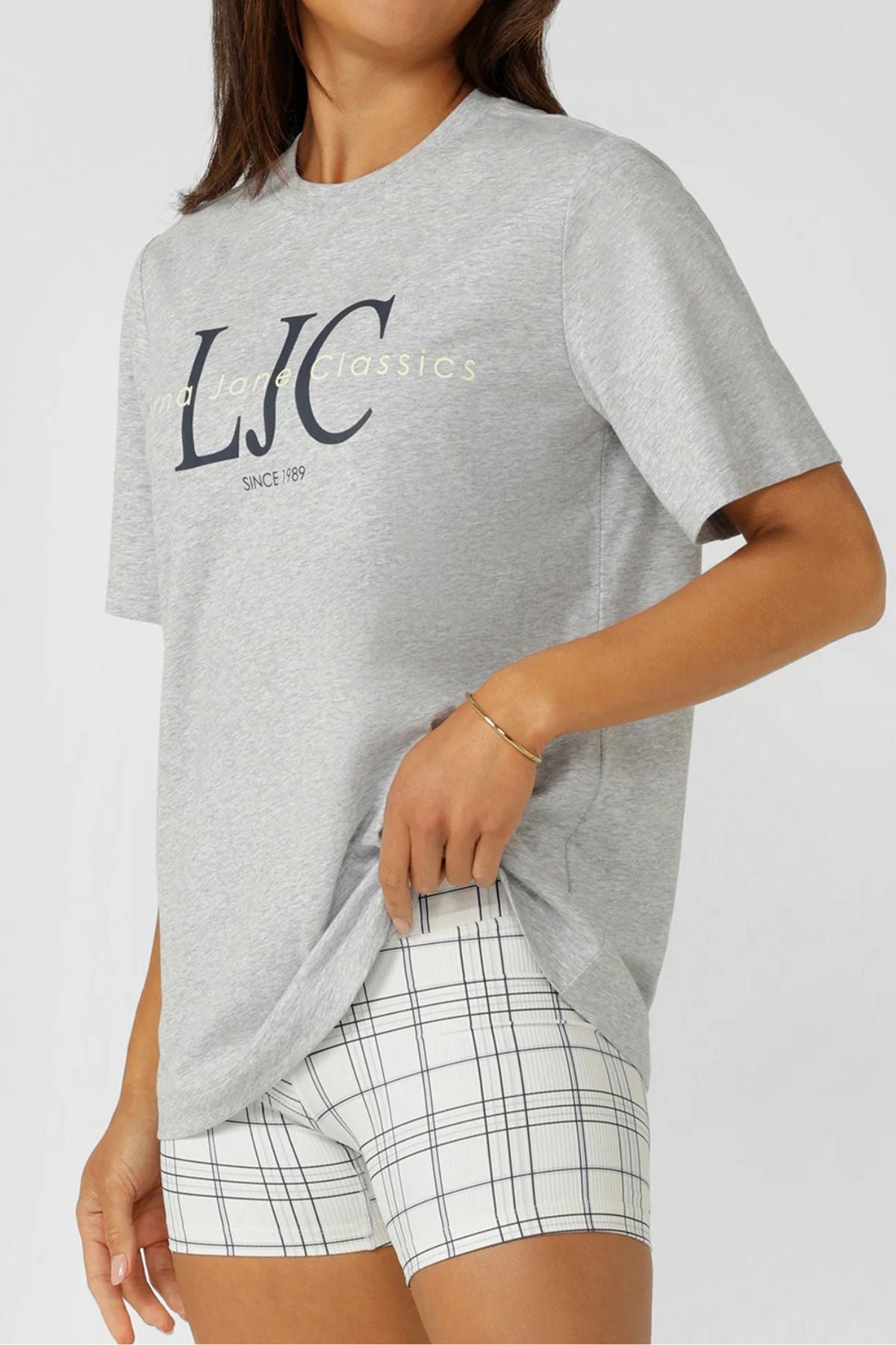 Wearing the Lorna Jane Heritage Classics Relaxed T-Shirt in grey, paired with white plaid shorts, a person stands against a plain light background, holding the hem of their t-shirt with one hand.