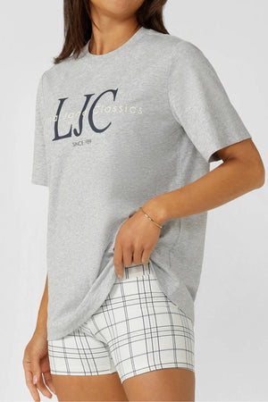 Wearing the Lorna Jane Heritage Classics Relaxed T-Shirt in grey, paired with white plaid shorts, a person stands against a plain light background, holding the hem of their t-shirt with one hand.