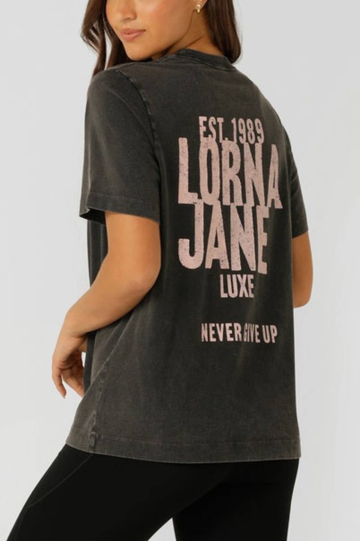 A woman wears the Lorna Jane Heritage Inspiration Relaxed T-Shirt in a vintage wash with an oversized fit and pink “EST. 1989 LORNA JANE LUXE NEVER GIVE UP” text on the back, standing sideways and looking over her shoulder.