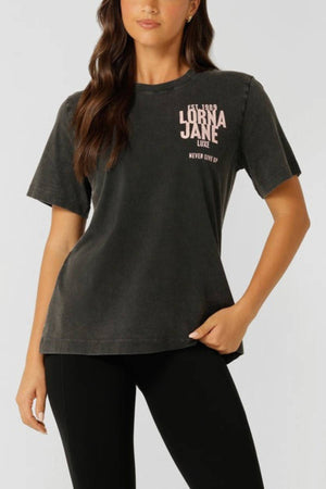 A woman wears the Lorna Jane Heritage Inspiration Relaxed T-Shirt in a vintage wash with "Lorna Jane Luxe" and "Never Give Up" in pink, styled oversized with black leggings, standing against a plain background.