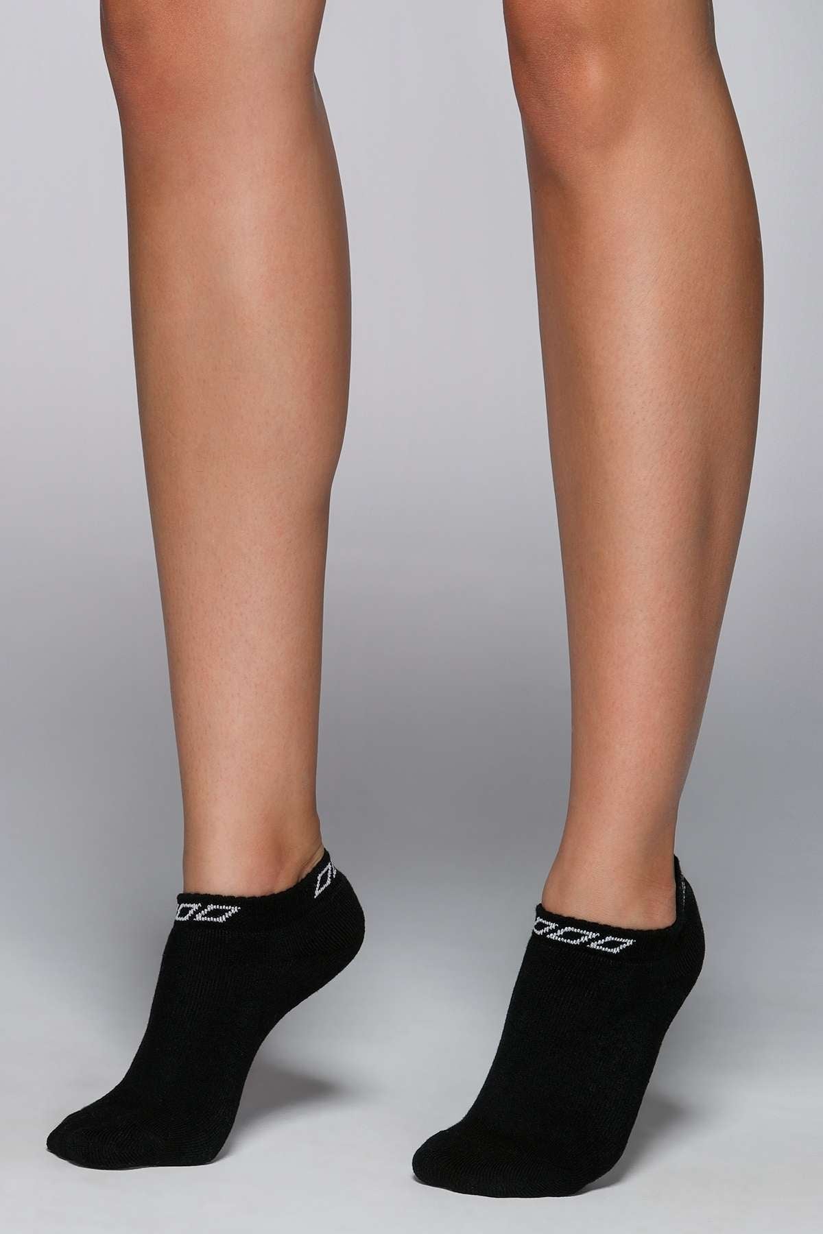 Someone is wearing Lorna Jane Iconic Socks—black ankle-length socks featuring a white geometric pattern near the cuffs—while standing on a light gray surface. Only the lower legs and feet are shown, showcasing their comfort and breathability.