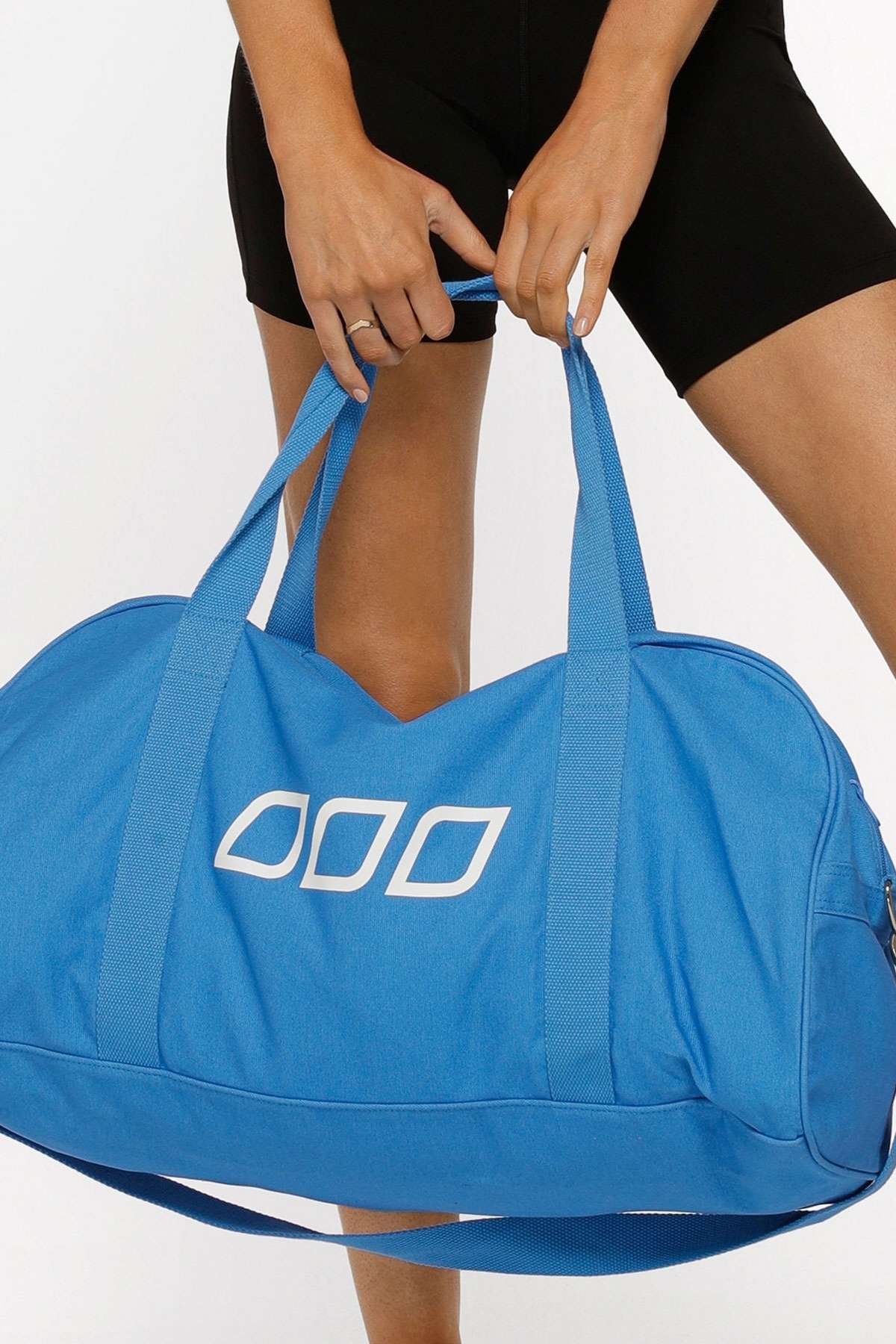 A person in black shorts holds the Lorna Jane Icons Canvas Duffle Bag, a large blue bag with white geometric patterns and an adjustable strap, standing against a plain white background.