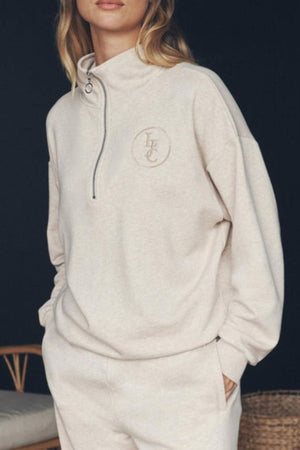 A woman stands against a dark background wearing the Lorna Jane LJC Heritage Quarter Zip Sweat in light beige with a small embroidered chest logo, matching sweatpants, and her hand in her pocket.