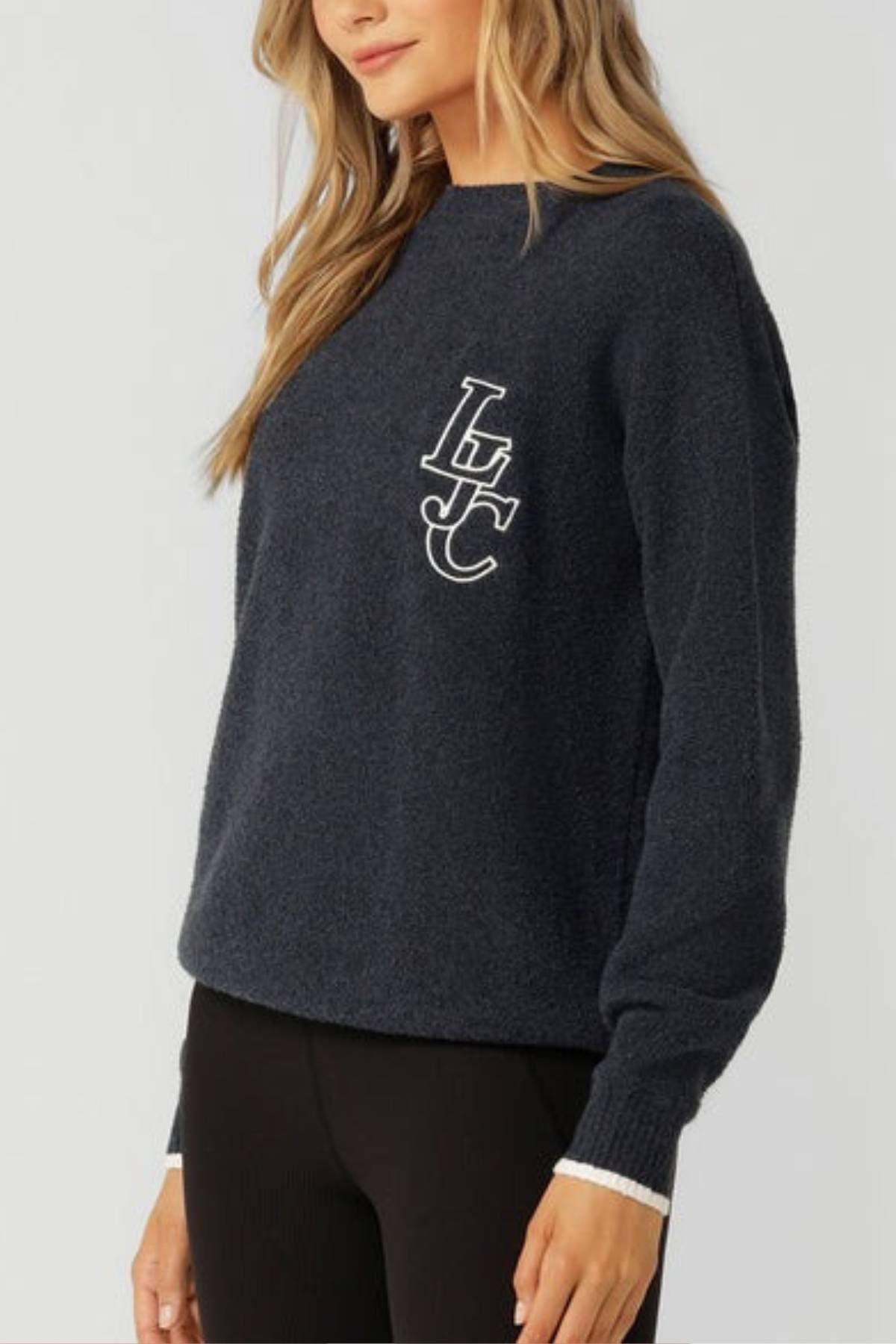 A woman with long blonde hair wears the Lorna Jane LJC Boucle Knit Jumper in dark navy, featuring white "LFC" letters on the chest, paired with black pants. The image is cropped from shoulders down against a plain light background.