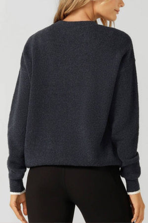 A woman with long blonde hair faces away, wearing a dark gray Lorna Jane LJC Boucle Knit Jumper and black pants. She is seen from shoulders to upper thighs against a plain light background.