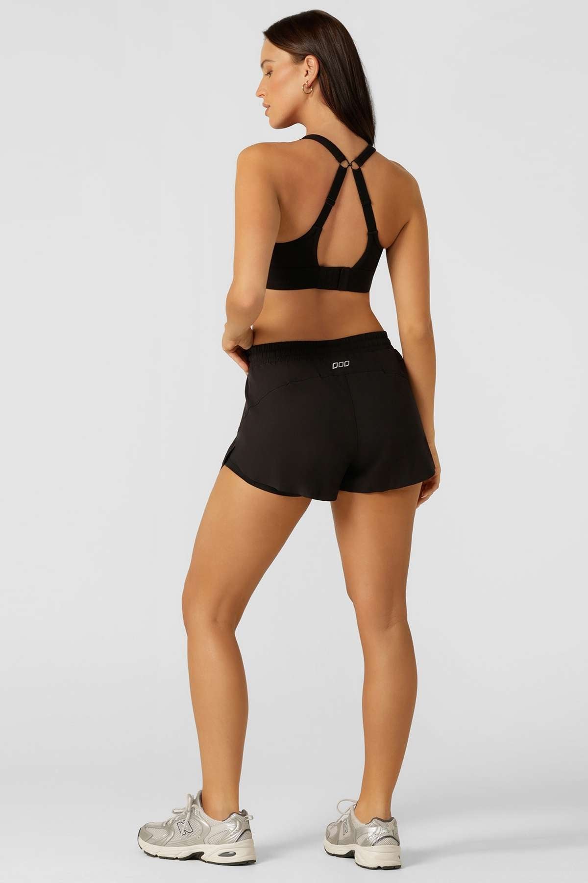 A woman stands facing away from the camera, wearing a black sports bra with crisscross straps, Lorna Jane Lightspeed Phone Pocket Run Shorts in black, and white sneakers, set against a plain light background.