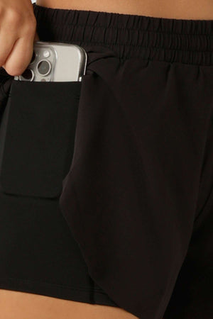 A person places a smartphone into the internal pocket of black Lorna Jane Lightspeed Phone Pocket Run Shorts made from sweat-wicking fabric.