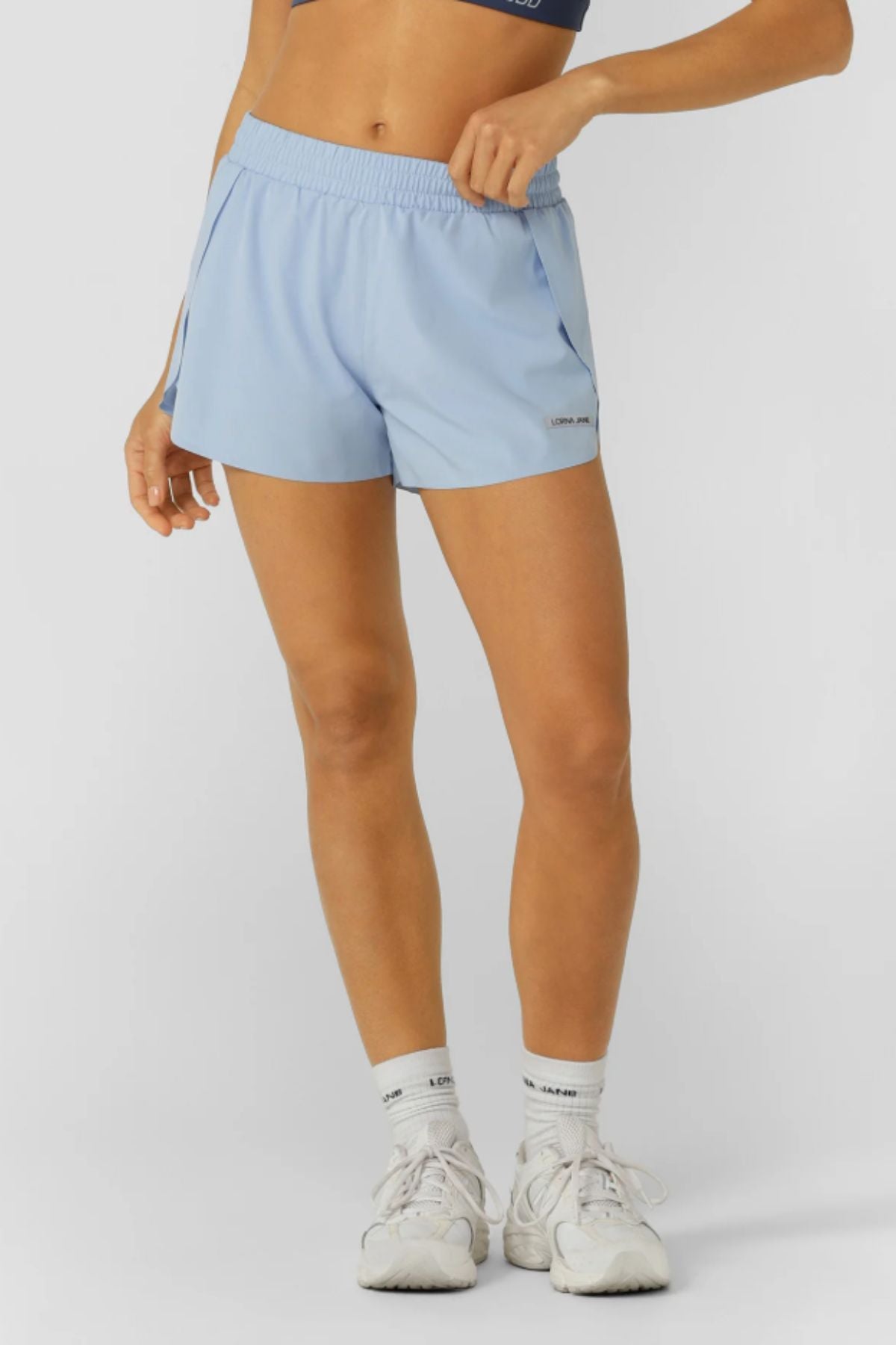 Wearing Lorna Jane Lightspeed Phone Pocket Run Shorts in light blue, a dark crop top, white socks, and white sneakers, a person stands against a plain light background with only the lower torso and legs visible.