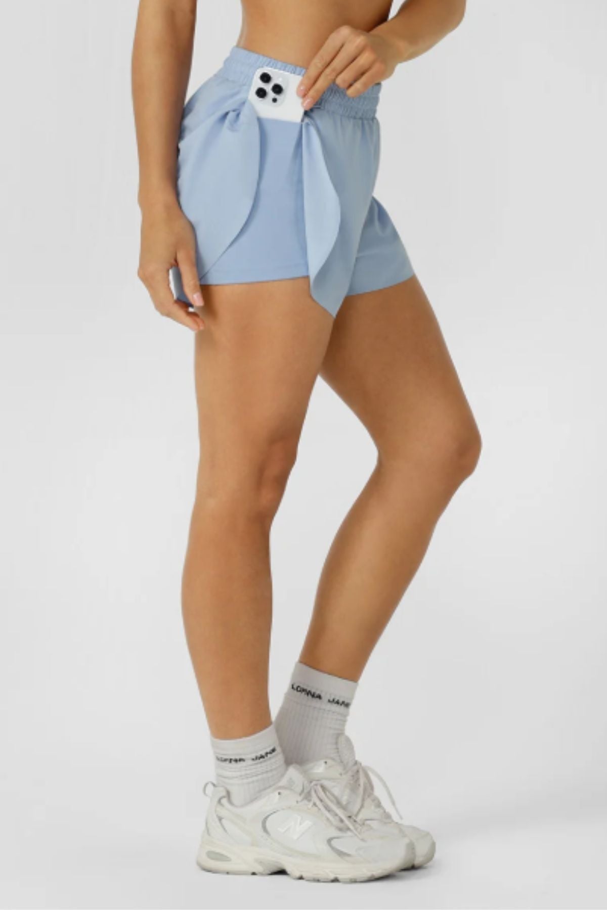 A person wears Lorna Jane Lightspeed Phone Pocket Run Shorts in light blue, with a phone in the pocket, paired with white socks and athletic shoes, standing against a plain light background.