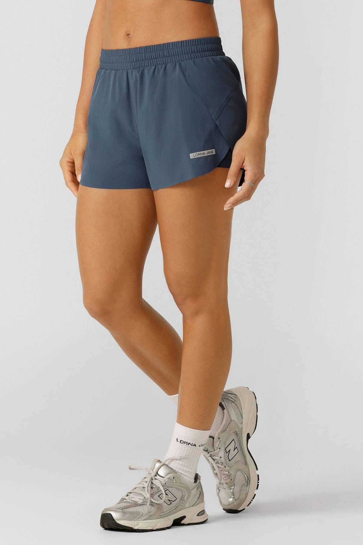 A person wearing blue Lorna Jane Lightspeed Phone Pocket Run Shorts, a matching sports top, white socks, and gray New Balance sneakers stands against a plain light background. Only the torso and legs are shown.