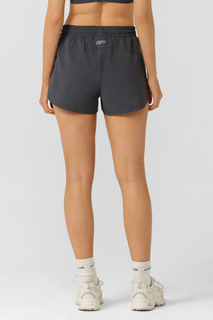 Someone wearing a black sports bra, Lorna Jane Lightspeed Phone Pocket Run Short, white crew socks, and white sneakers stands with their back to the camera against a plain light gray background.