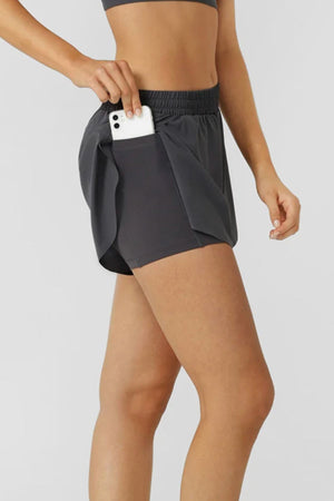 A person wearing Lorna Jane Lightspeed Phone Pocket Run Shorts in dark gray slips a white smartphone into the side pocket. Shown waist to thigh against a plain light background, the shorts have sweat-wicking fabric for comfort.