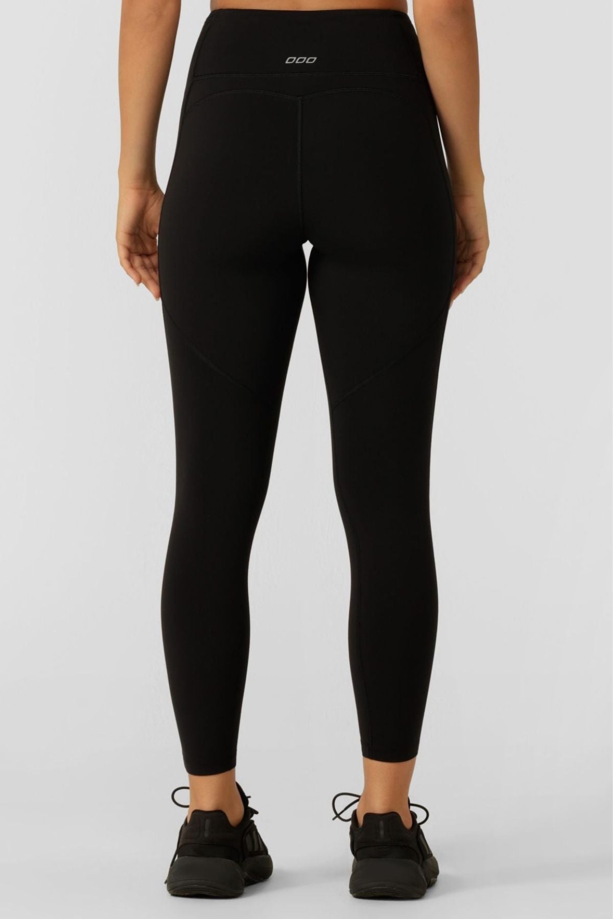 A person stands with their back to the camera, wearing Lorna Jane Amy Thermal Phone Pocket Ankle Biter Leggings and black athletic shoes against a plain light background.