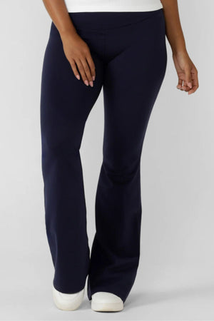 A person wearing Lorna Jane Lotus Flared Legging in navy blue and white sneakers stands against a plain light background, showing only the lower torso, legs, and one arm.