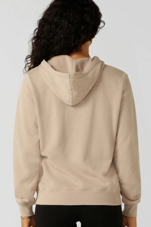 Seen from behind, a person with long curly hair wears the Lorna Jane Lotus Hoodie in beige and black pants, standing against a plain light background.