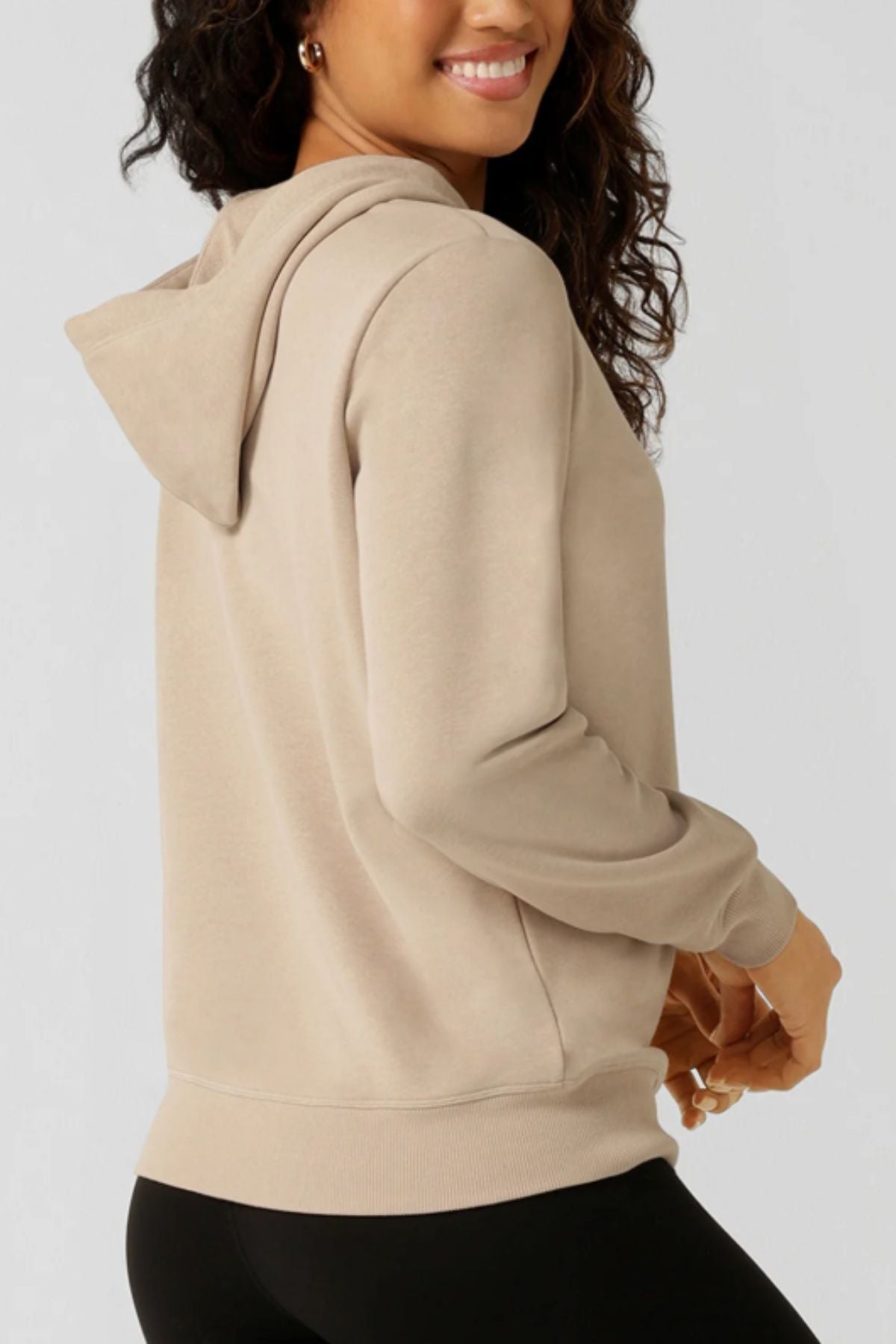 A woman smiles and turns to the side, wearing the Lorna Jane Lotus Hoodie in light beige with black pants, set against a plain light gray background. The Lotus Hoodie is lightweight—a perfect pick for workouts or casual days.