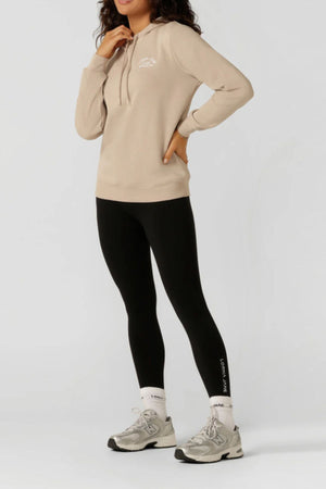 Against a plain background, a person in a beige Lorna Jane Lotus Hoodie, black leggings, white socks, and gray sneakers poses with their left hand on their hip; their head is partially out of frame.