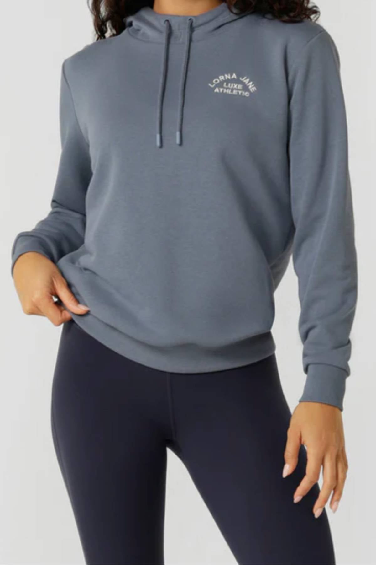A woman stands against a light background, wearing the Lorna Jane Lotus Hoodie—a cozy cotton blend with “Lorna Jane Luxe Athletic” on the chest—and black leggings, one hand resting on her hip.