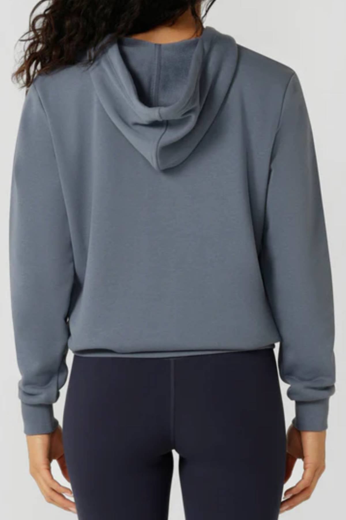 A person with long, wavy dark hair is seen from behind, relaxed in the gray Lorna Jane Lotus Hoodie and dark leggings—a classic women’s activewear look with the hood down and arms at their sides.