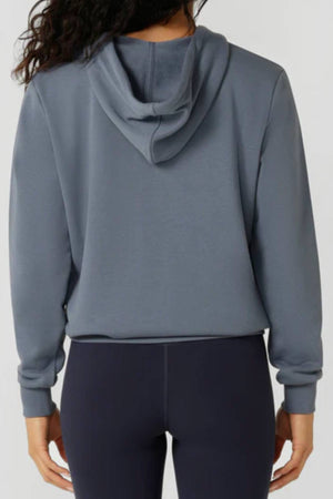 A person with long, wavy dark hair is seen from behind, relaxed in the gray Lorna Jane Lotus Hoodie and dark leggings—a classic women’s activewear look with the hood down and arms at their sides.