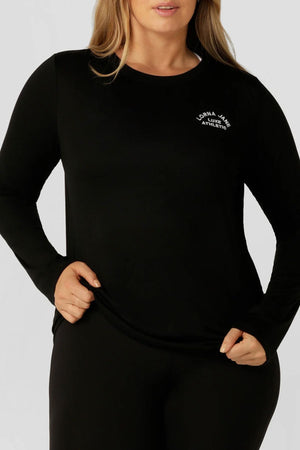 A woman stands against a neutral background wearing a fitted Lorna Jane Lotus Long Sleeve Top with a small white "Lorna Jane Athletics" logo, paired with sleek black pants.