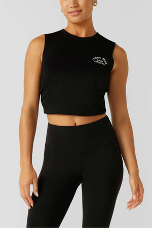 A woman stands against a light gray background wearing the Lorna Jane Lotus Muscle Tank, a black sleeveless crop top with white text, paired with high-waisted black leggings for lightweight comfort and easy layering.
