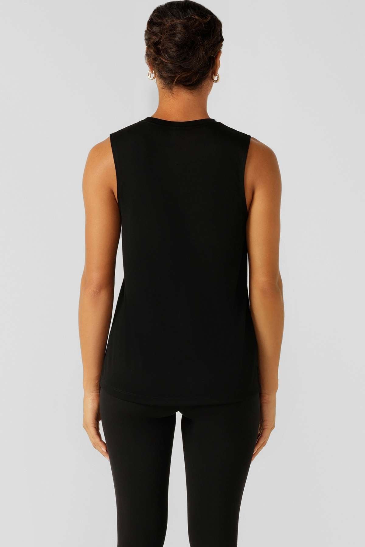 A person with short, dark hair, wearing the Lorna Jane Lotus Muscle Tank and black pants, stands facing away from the camera against a plain light background, highlighting the tank's relaxed silhouette.