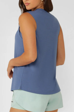 A woman with brown hair down, facing away and looking over her shoulder, wears a sleeveless blue Lorna Jane Lotus Muscle Tank and light green shorts, standing against a plain light background.