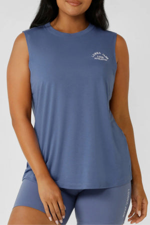 A woman wears the Lorna Jane Lotus Muscle Tank, a lightweight, breathable top perfect for layering, as she poses with one hand by her side.