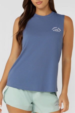A woman wears a sleeveless blue Lorna Jane Lotus Muscle Tank with “Lorna Jane Luxe Athletic” text on the chest and light green shorts, standing against a plain light background.