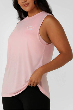 A woman stands in a sleeveless light pink Lorna Jane Lotus Muscle Tank featuring a small white logo on the left chest, paired with black leggings and holding the hem of her breathable top.