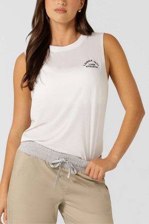 A woman wears the Lorna Jane Lotus Muscle Tank, a sleeveless white top with a heritage embroidered logo, paired with light beige drawstring pants. The breathable natural blend makes this lightweight look perfect for layering.