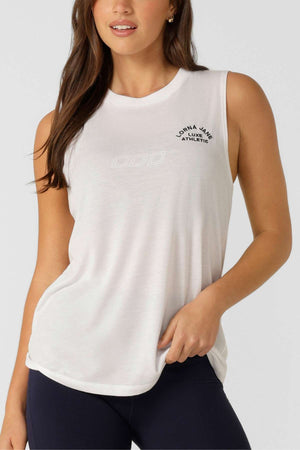 A woman wears the Lorna Jane Lotus Muscle Tank, a sleeveless white athletic top with a heritage embroidered logo on the left chest, paired with dark leggings. She poses holding the hem of her tank against a plain background.