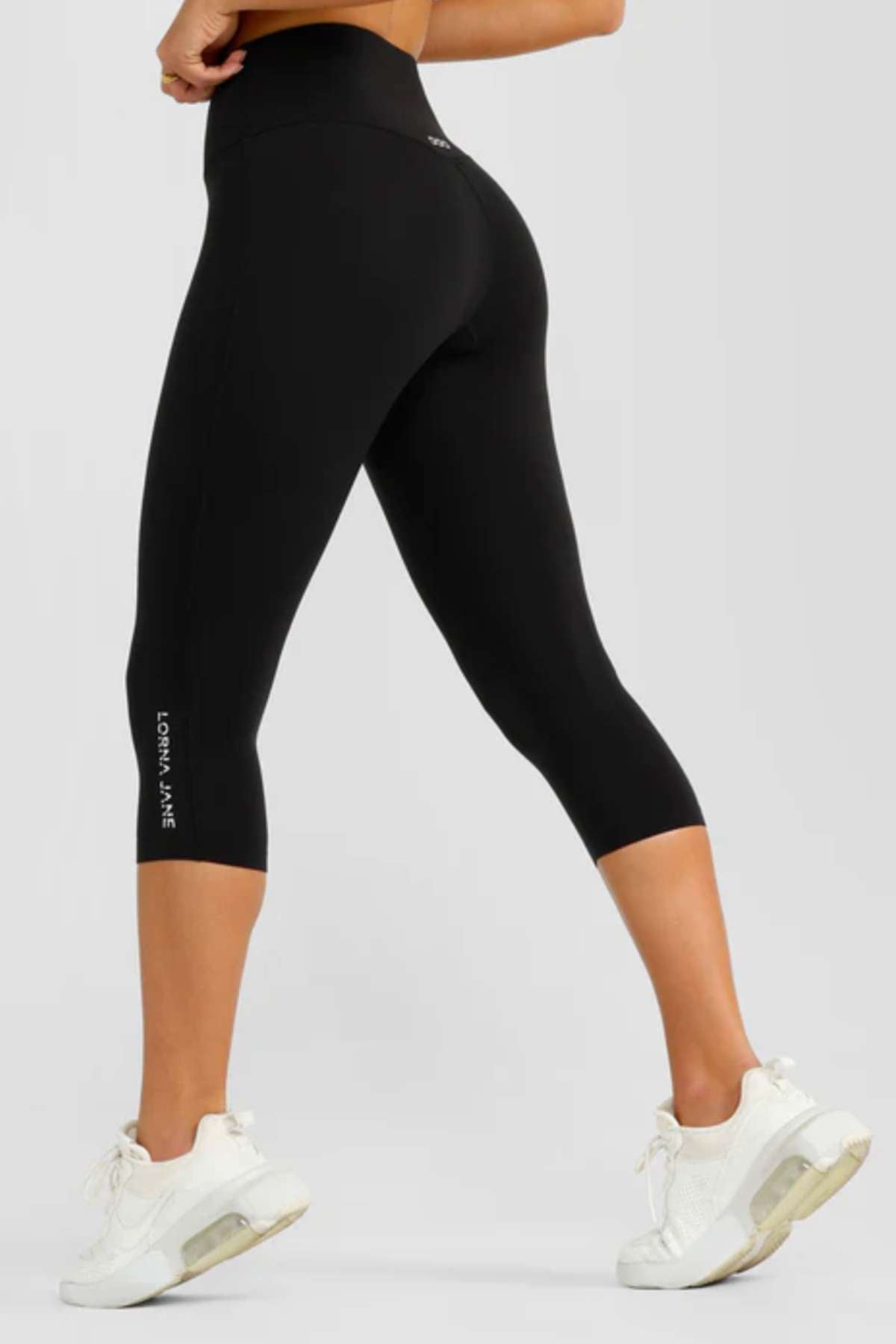 A person stands against a plain light background wearing black Lorna Jane Lotus No Chafe 7/8 Leggings with a high-rise fit and white sneakers. The leggings display "LORNA JANE" along the left calf.