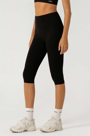 A person in a black sports bra and Lorna Jane Lotus No Chafe 7/8 Leggings stands against a plain light background. These high-rise, moisture-wicking leggings are ideal for active days, paired with white socks and athletic shoes.