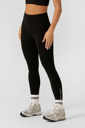 A person wears Lorna Jane Lotus No Chafe Ankle Biter Leggings in black, paired with a black crop top, white socks, and gray running shoes, standing against a plain light gray background. Their head and feet are out of frame.